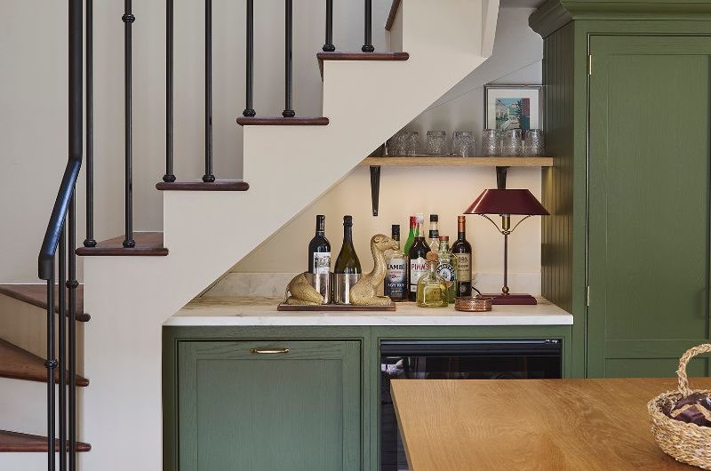 A home bar design with green cabinets, wine fridge, small countertop space and a compact shelf, under the stairs.