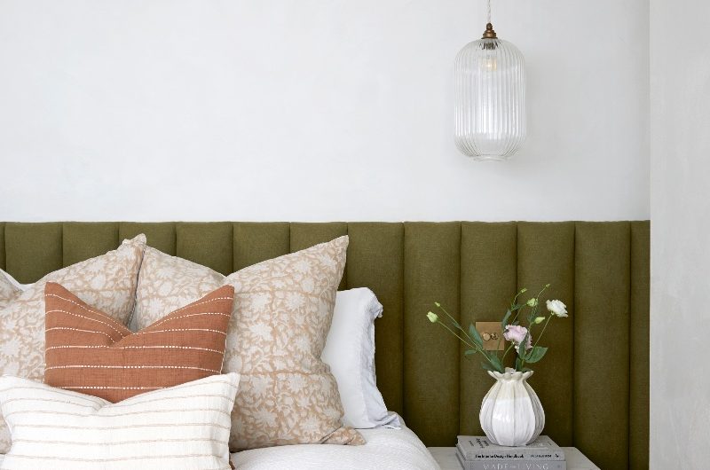 A neutral bedroom with a green upholstered bed frame, white bed linen, a wood bedside table, and a pendant light with fluted glass.