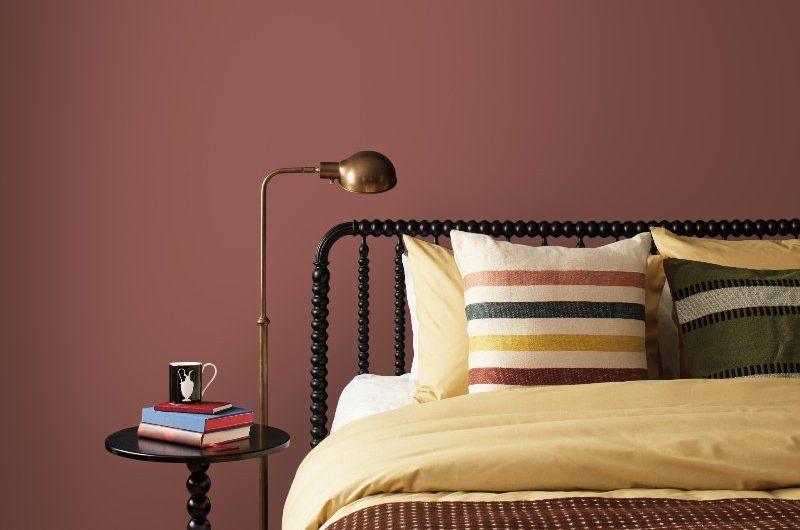 A neutral bedroom design with dark brown wall paint, bobbin bed frame and matching side table, mustard yellow bed linen, brass tap, and patterned cushions.