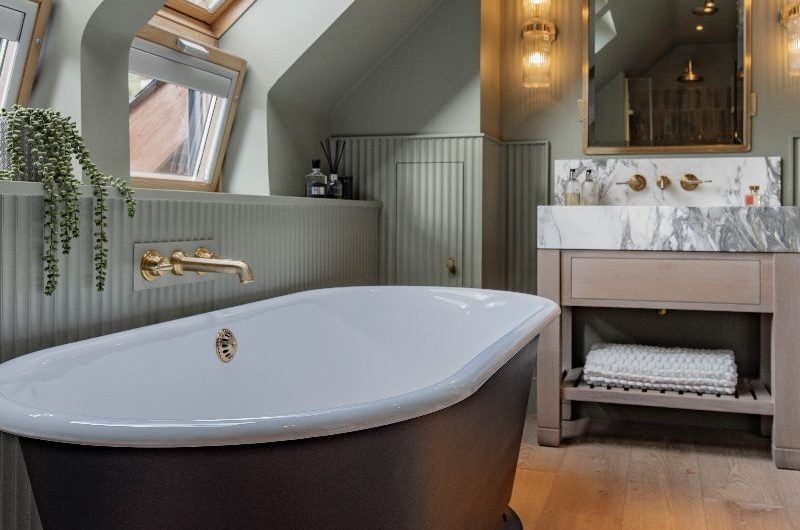 A bathroom renovation project with sage green wall paint, a freestanding tub, a wood and stone vanity and brass details.