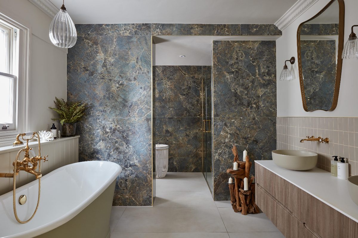 A large primary ensuite with stone-effect tiles on a partition wall separating the shower, with a wood double vanity, and a freestanding tub.