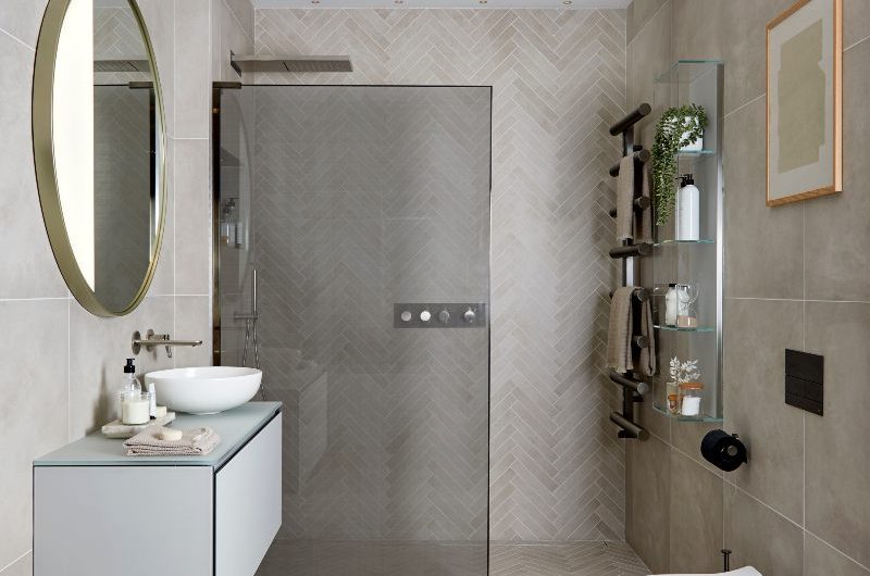 A neutral wetroom with herringbone and large-format tiles, a towel rail, wall-mounted storage, and a contemporary vanity with a gold circular mirror.