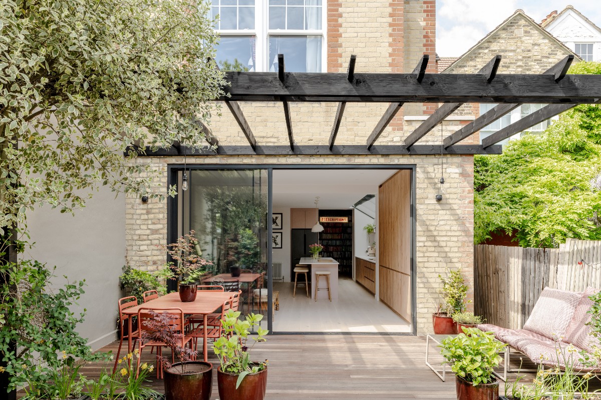 View inside a kitchen-diner from a patio with colourful garden furniture, greenery, and a wood pergola.