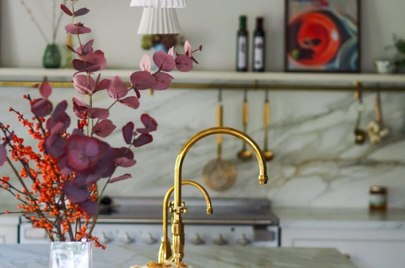 A calm kitchen design with veiny worktops, an island with a sink and brass taps, and colourful decor.