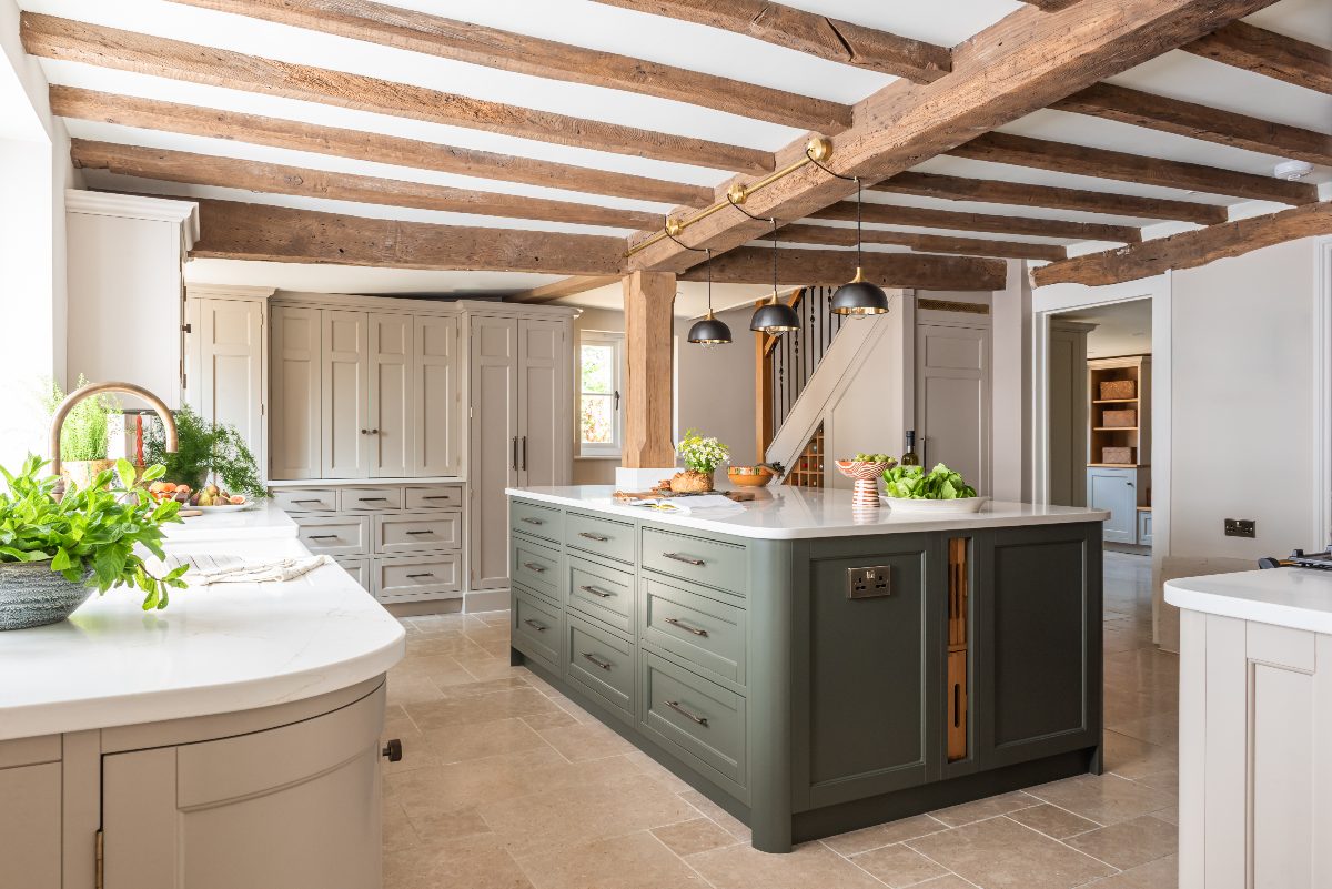 Classic kitchen design with cream Shaker country and a dark green island with storage, power sockets, ample worktop space and pendant lights hanging from wooden beams across the ceiling.