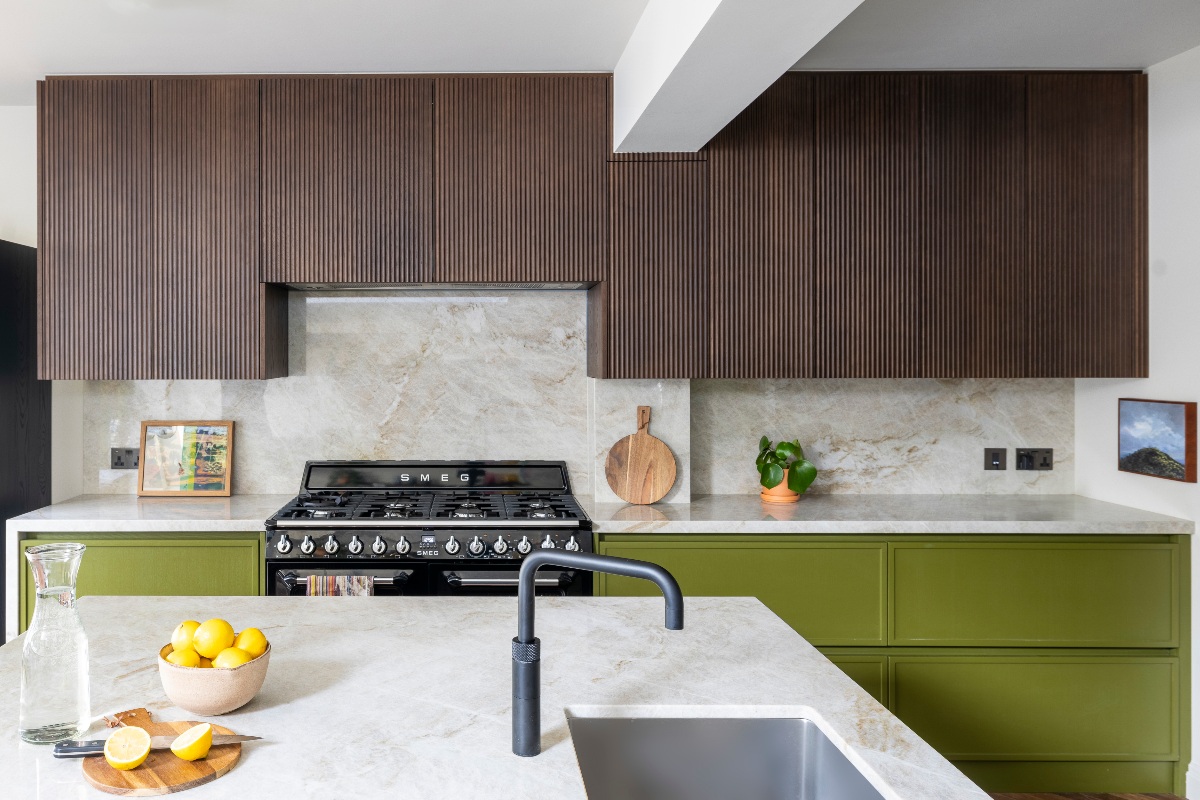 A cooking run with green base units, fluted wood wall units, veined splashback and work surfaces, a black range cooker with concealed extraction, and a central kitchen island.