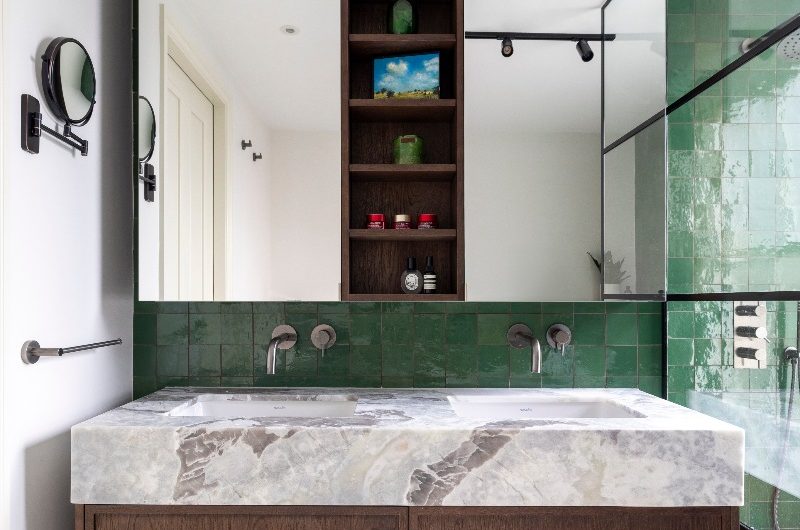 A vanity area with a wall-mounted wood unit with statement stone and two inset basins, with green zellige tiles on the wall and a double mirror shelving unit above it.