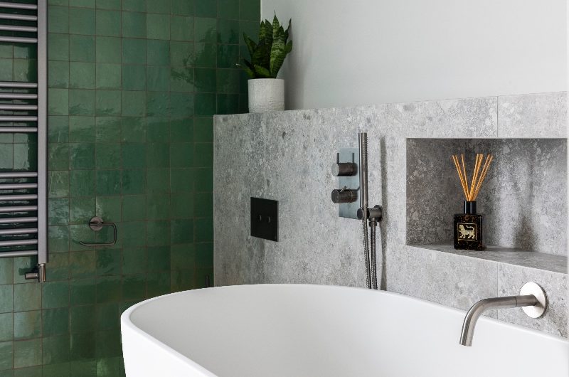 A bath zone with a spacious white tub, chrome fittings, a stone-clad half wall with a storage niche behind it, and green zellige tiles on the opposite wall.