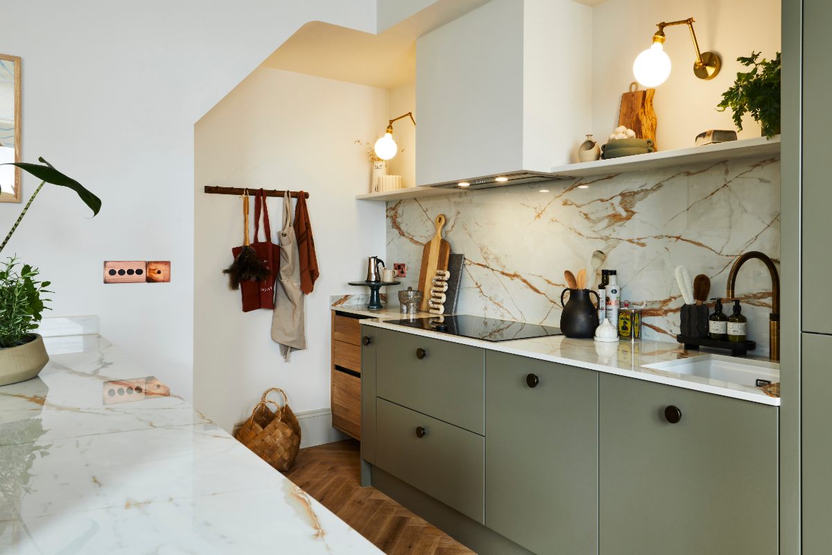A green kitchen design with gold and white veiny worktops, round black handles, layered lighting, and concealed extractor above the induction hob.