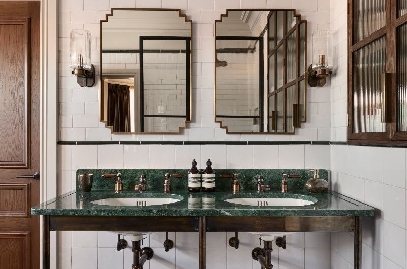 A boutique hotel style bathroom with patterned flooring, a double basin unit with green marble, Art Deco mirrors and fluted wall lights.