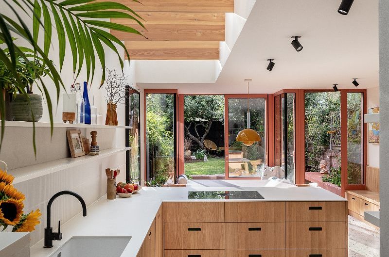 A modern kitchen design with L-shaped wood cabinetry with speckled terrazzo flooring, a wide rooflight with oversized timber louvres, and peachy glazing.