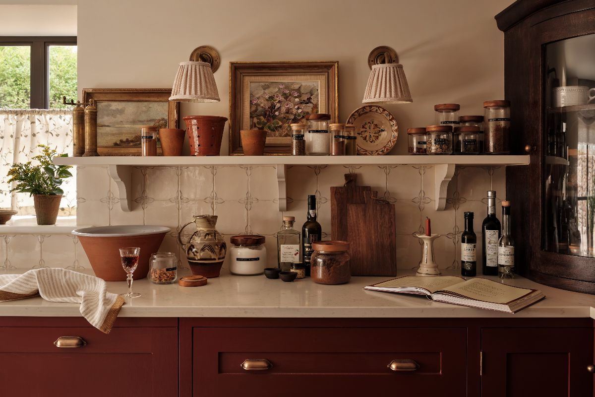 A country kitchen with burgundy red cabinetry, white work surfaces, Delft splashback tiles, and open shelving with rustic decor.