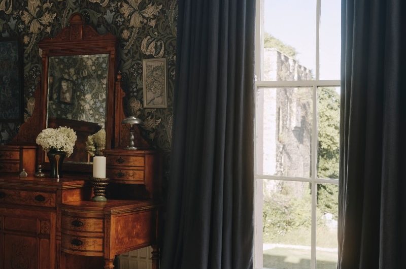 A bedroom design with animal print carpet, floral wallpaper, chunky blue curtains, and a vintage wood vanity.
