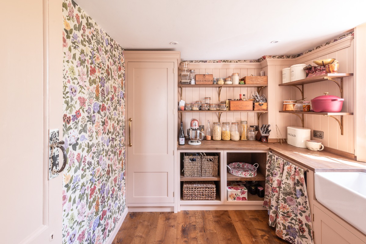 A pantry with pink kitchen cabinets, extra worktop space, a sink and tap, open shelving, and floral wallpaper.