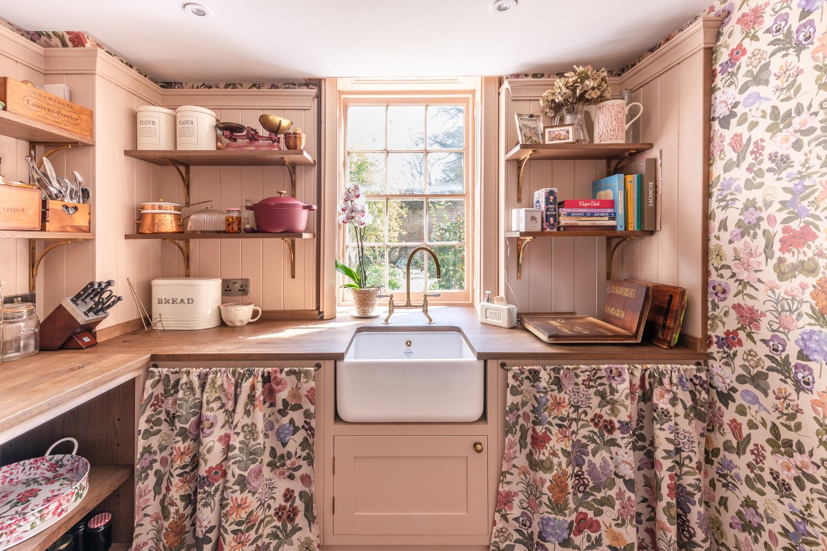 A walk-in pantry with wood worktops, a small butler sink and tap, classic panelling painted in pink, and floral wallpaper and sink skirts.