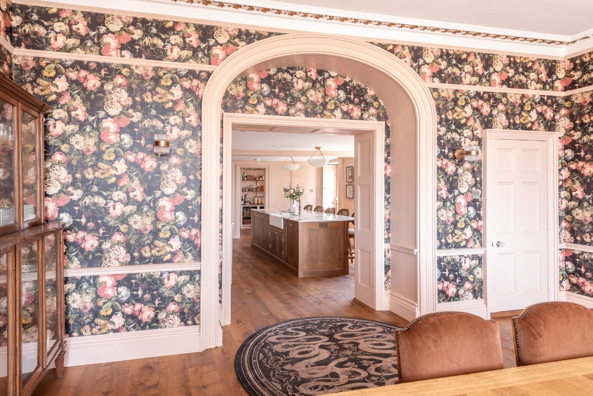 A romantic dining room design with statement floral wallpaper looking into the kitchen.