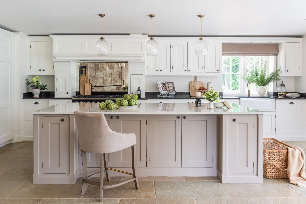 A traditional scheme with white cabinetry and contrasting black worktops, an island in a warm taupe colour, with white worktops and seating, and stone flooring.