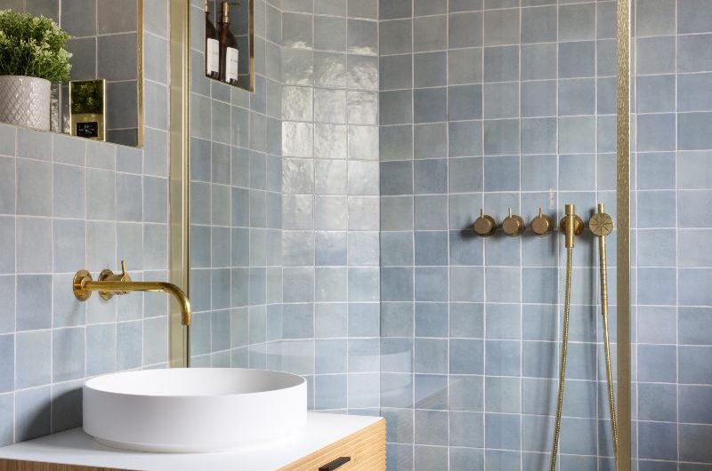 A closeup at a walk-in shower with blue square wall tiles, gold panel and shower system, and a wood vanity next to it.