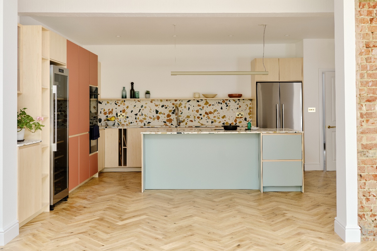 A retro kitchen with handleless cabinetry in wood, terracotta and light blue colours, with terrazzo worksurfaces, and herringbone flooring.