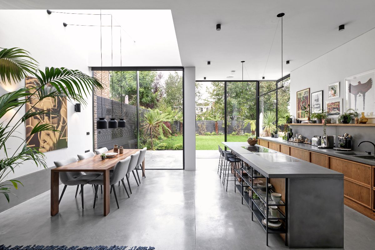 Industrial scheme with wood units, concrete-effect worktops and matching island, with open steel shelving, a dining zone, and tall and wide glazing.