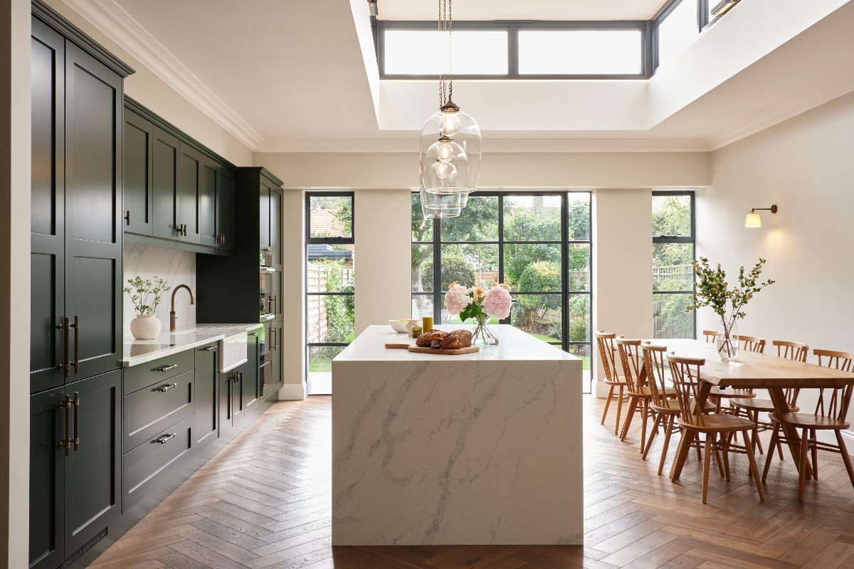 A spacious cooking and dining scheme with dark green cabinetry, a central stone-clad island, a wood table and chair set, and steel-framed glazing.