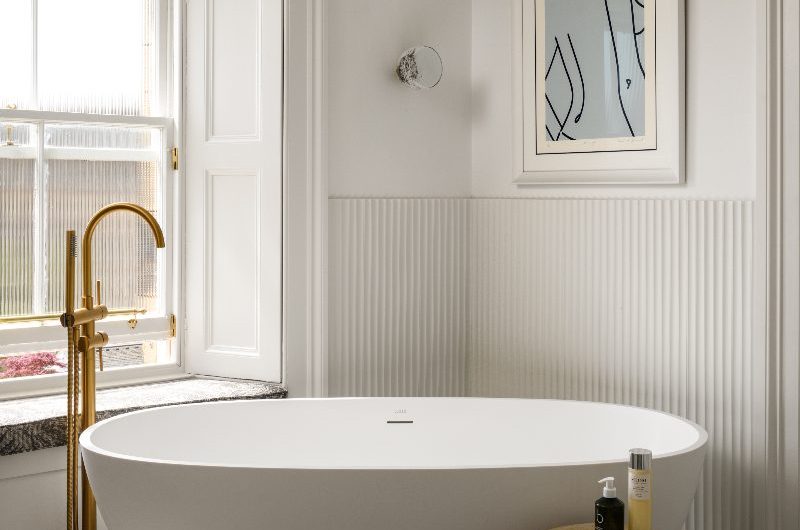 A modern bathroom zone with marble flooring, fluted wall panelling, a freestanding tub with a gold bath filler, and a wood stool for accessories.