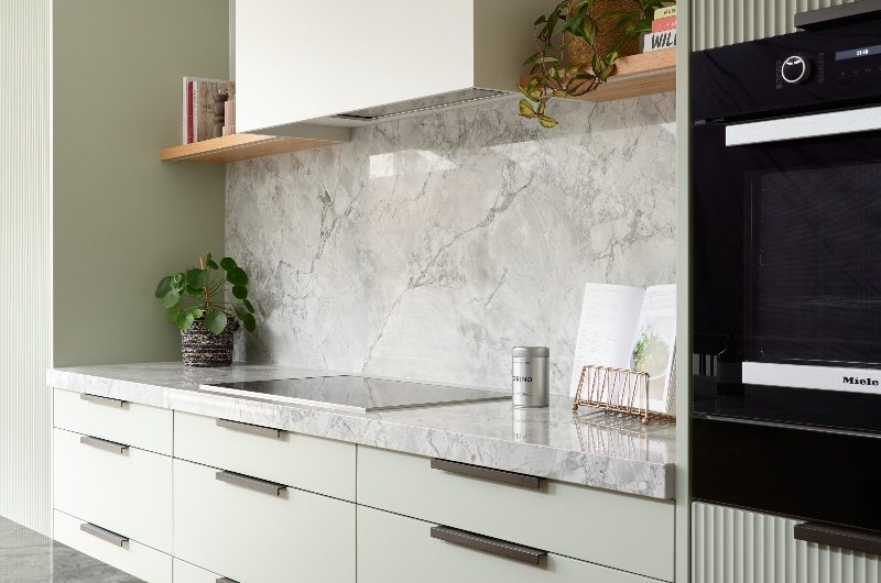 A modern fusion kitchen with sage green units with veiny worksurfaces, wood open shelving, and a concealed extractor.