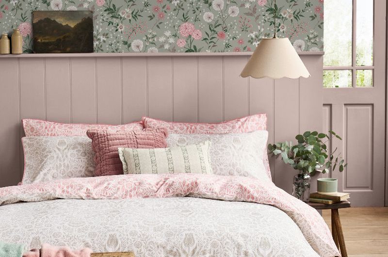 A country-chic bedroom with dusty pink wall panelling, floral wallpaper on a light green background, and a bed layered with cushions and throws.