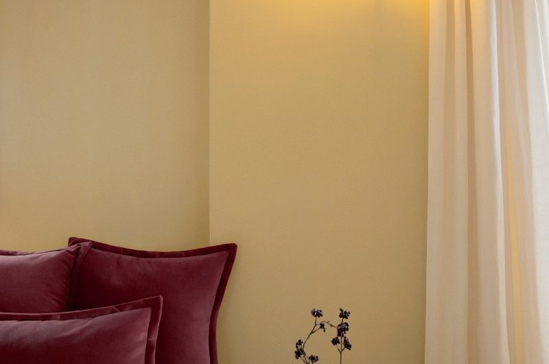A neutral bedroom with airy curtains, white bed linen, a rattan side table, and burgundy velvet cushions.