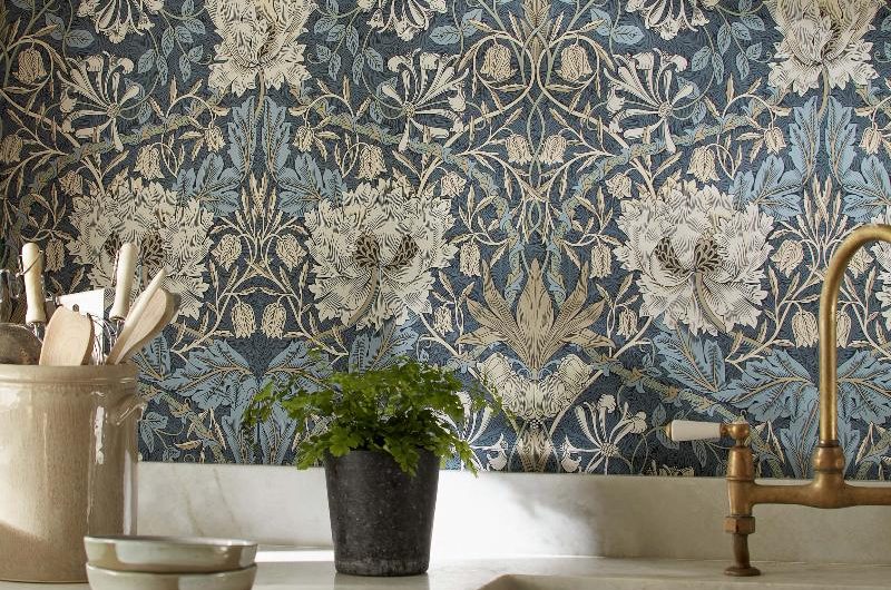 A closeup at a sink area with a butler bowl, brass tap, white worktops and floral wallpaper in the background.