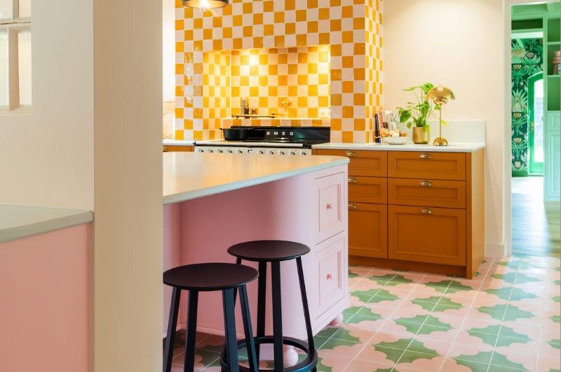 A colourful kitchen design with yellow base cabinets, a cooker zone with a concealed extractor drenched in checkerboard tiles, a pink island, and pink and green cement flooring.