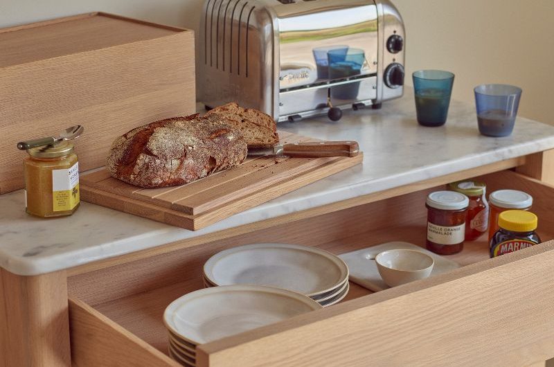 A freestanding wood breakfast unit with a chrome toaster and wood bread bin on the marble counter.