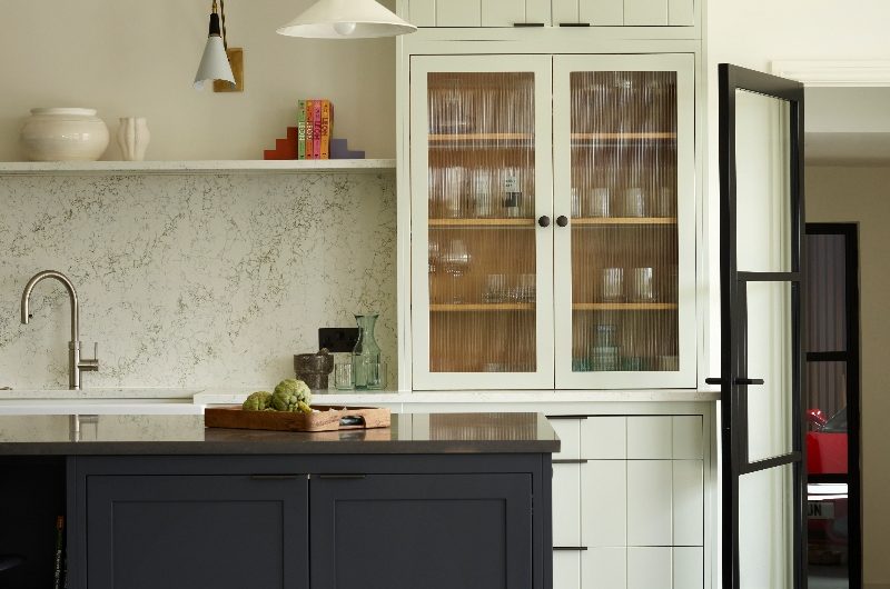 A main run of cabinets painted in an off-white colour with a mix of panelled doors and fluted glass, with a dark island at the heart of the design.