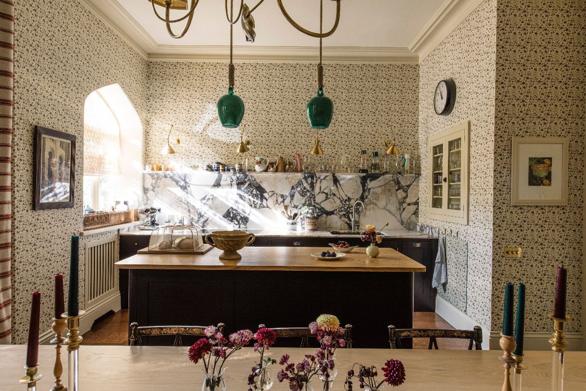 A bold kitchen design with patterned wallpaper, dark cabinetry and a matching island, with a mix of surfaces and decor.