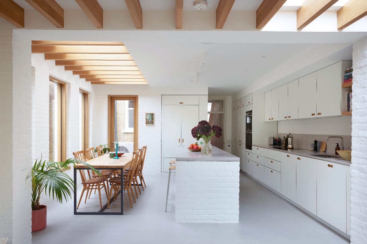 A white kitchen design with seamless cabinetry, cut-out handles, a central island, a dining zone under. a glass roof with timber beams, and minimalist finishes.