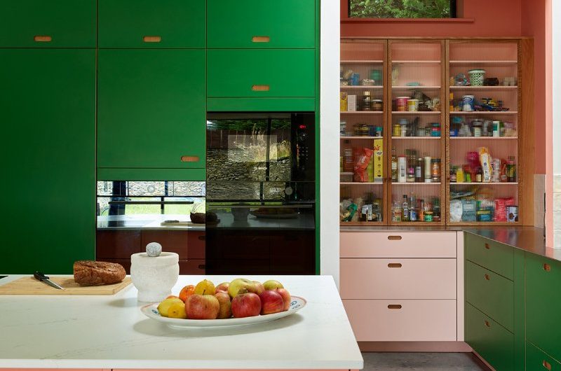 A three tone kitchen, with tall wall and base cabinets in rich green, an island in a colour shade, plus pale pink and reeded glass units in the open pantry.