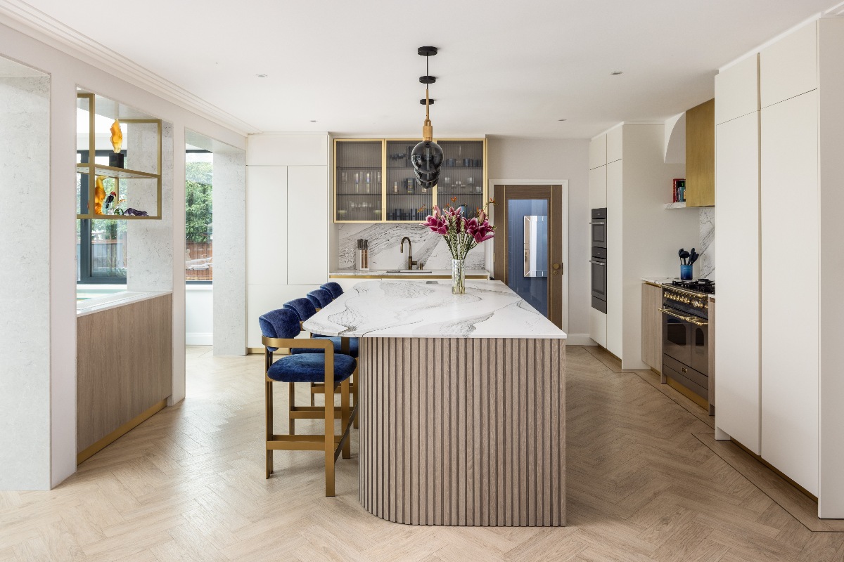 A contemporary scheme with white handleless cabinetry, brass and fluted glass wall-mounted units, a stainless steel range and brass extractor cover, and a central fluted wood island.
