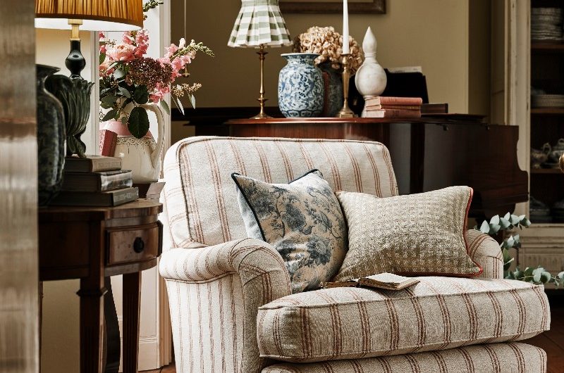 A rustic, country-style living room with wall art, a striped armchair, floral cushions, and classic table lamps on wood furniture.