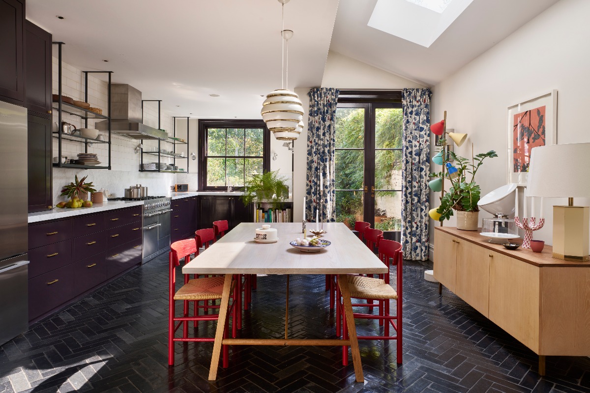A fusion kitchen with plum cabinetry, black herringbone tiles, a long white table with red chairs positioned in the centre of the room, and a blonde wood sideboard.