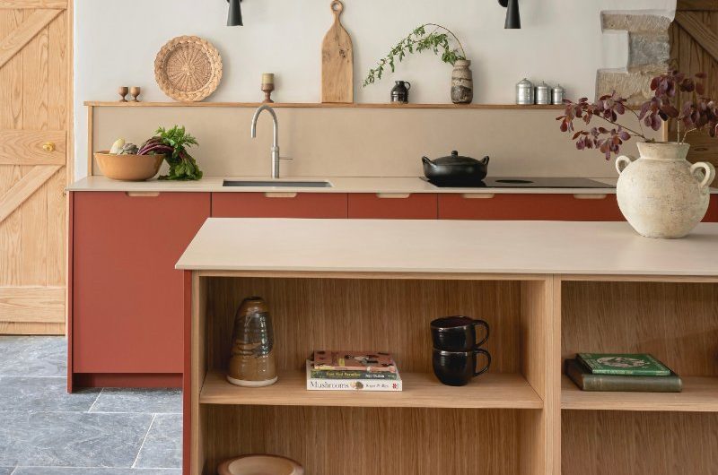 A rustic kitchen with dark red and plywood base cabinets and island, stone flooring, and sand-coloured work surfaces, and exposed beams.