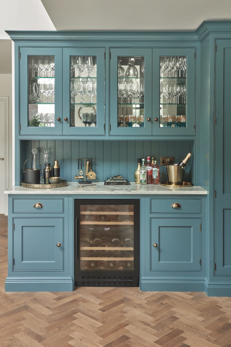 A blue home bar with glass wall cupboards, panelling, stone worktop, and a bottle fridge.