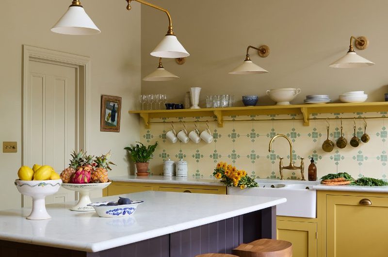 A colourful kitchen design with a dark brown-purple island, a run of units painted in yellow, with patterned tiles and open shelving.