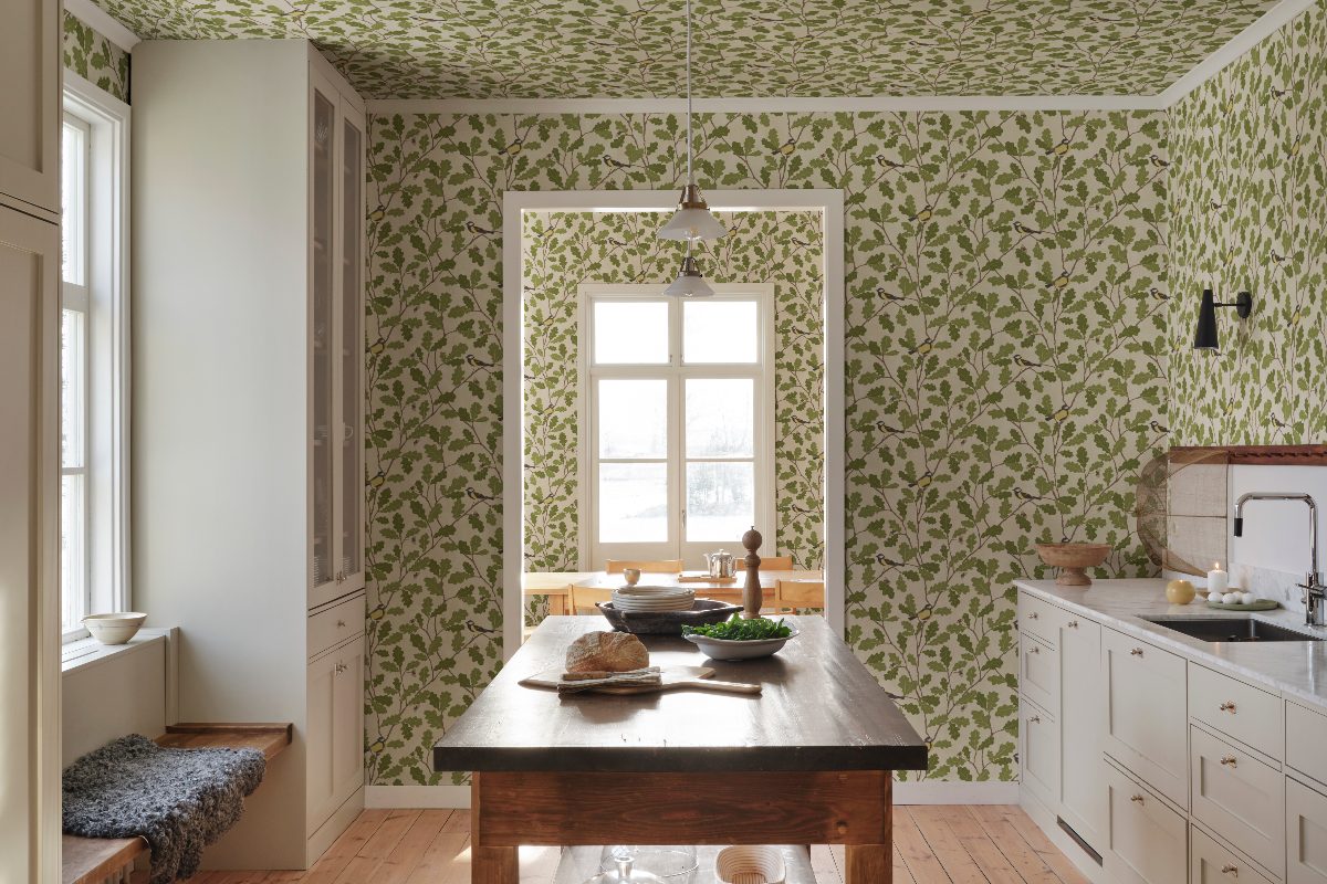 A white Shaker kitchen design with a main run of cabinets, a wood table in the centre, with nature-inspired green and white wallpaper on the walls, ceiling, and adjoining dining room walls.