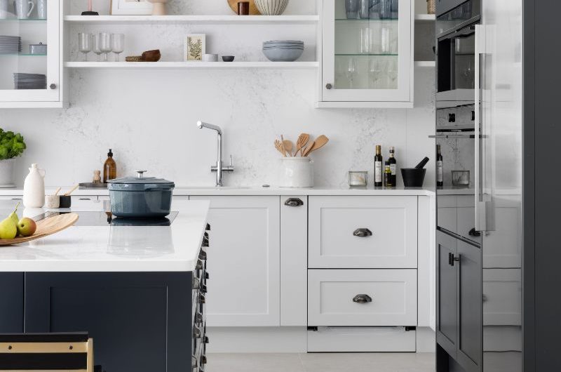 A white kitchen design with a blue island, open shelving and glass wall cabinets, with an appliance zone on the right, stone flooring, and underfloor heating.