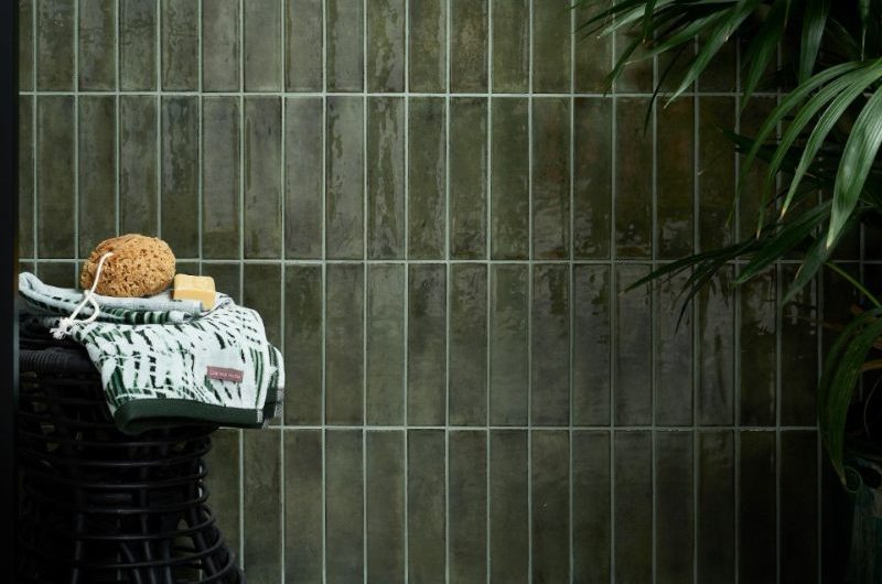 A shower zone with metro wall tiles in a dark green, with house plants, a stool for accessories, and veined green floor slabs.