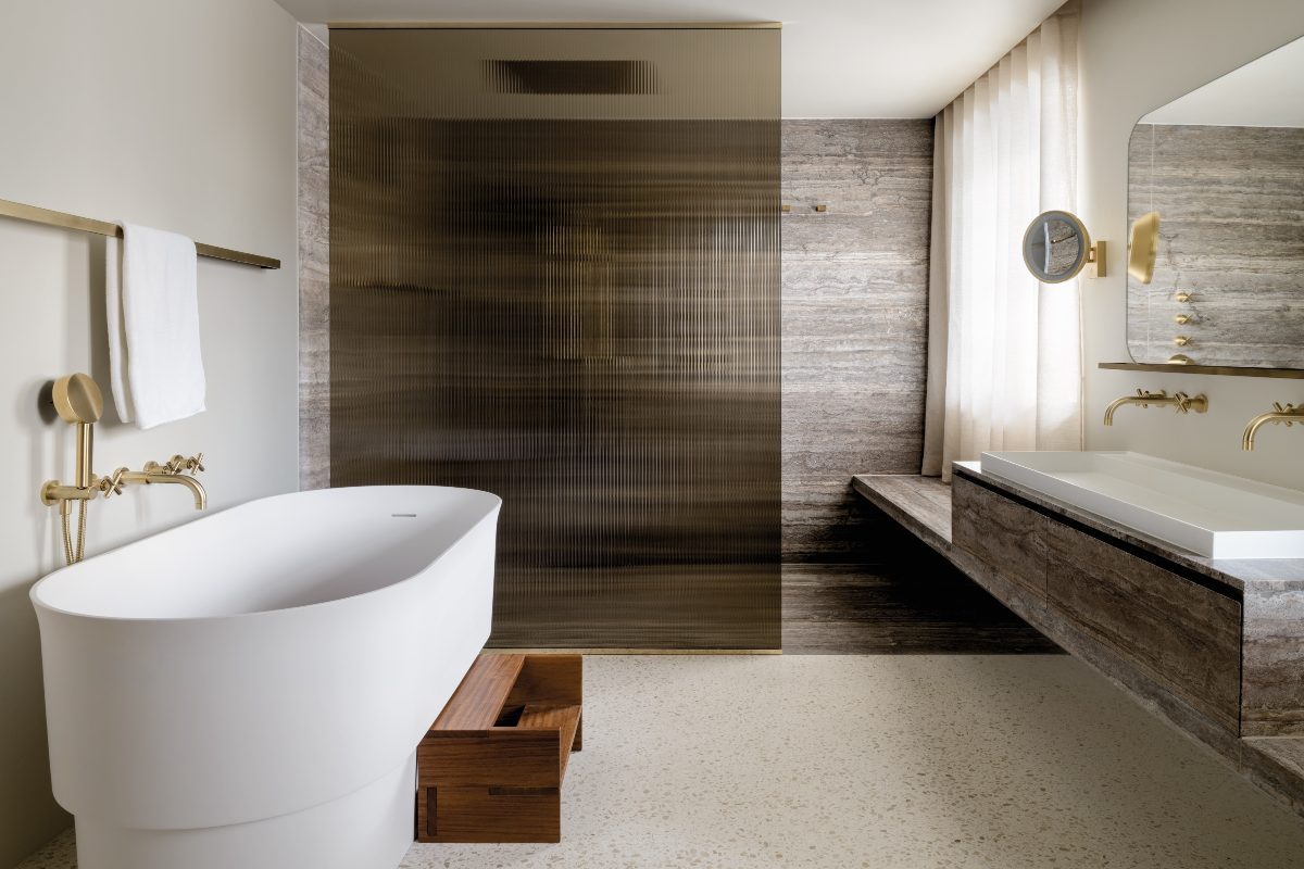 A modern scheme with terrazzo flooring, a darker shower area, a wall-mounted vanity with a long countertop basin, and a freestanding tub with gold brassware.
