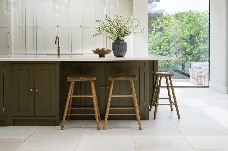 A classic scheme with timeless cabinetry, a green island with seating, breakfast bar and storage, seamless glazing, and stone-effect flooring.