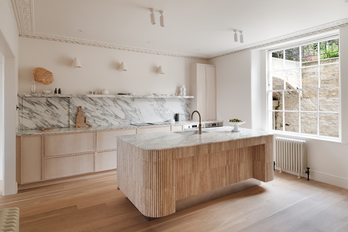 A neutral, minimalist kitchen design with a statement fluted travertine island with veiny Arabescato work surfaces, wood cabinetry, and wood flooring.