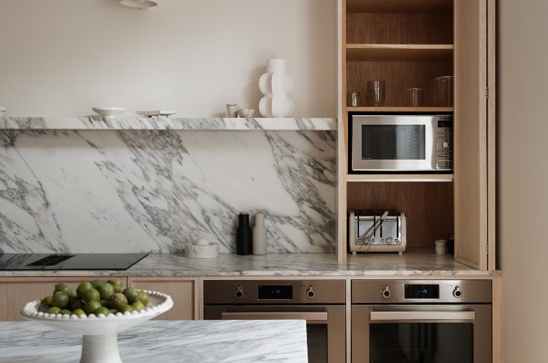 Closeup at an open dresser cupboard with bi-fold door which houses a microwave and toaster, with ovens built within the cabinetry underneath.