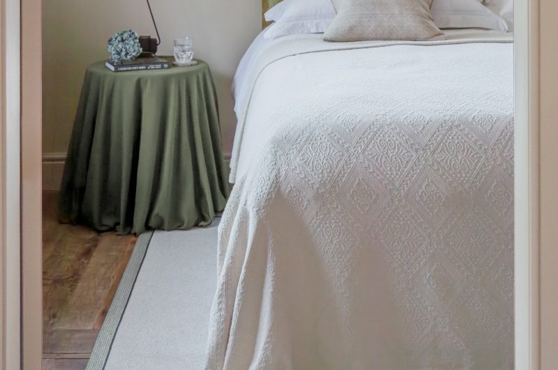 A bedroom design with white linen, a bedside table covered with a green fabric, and a boarded rug.
